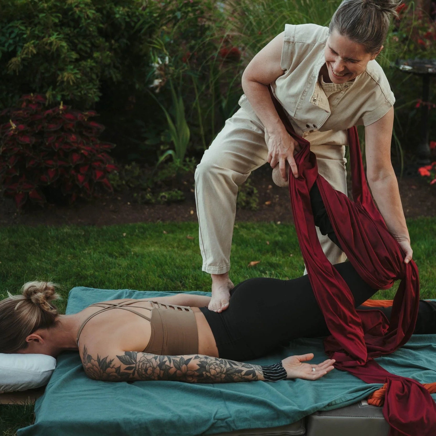Two women in a garden setting, one lying on a mat receiving a massage from another woman.