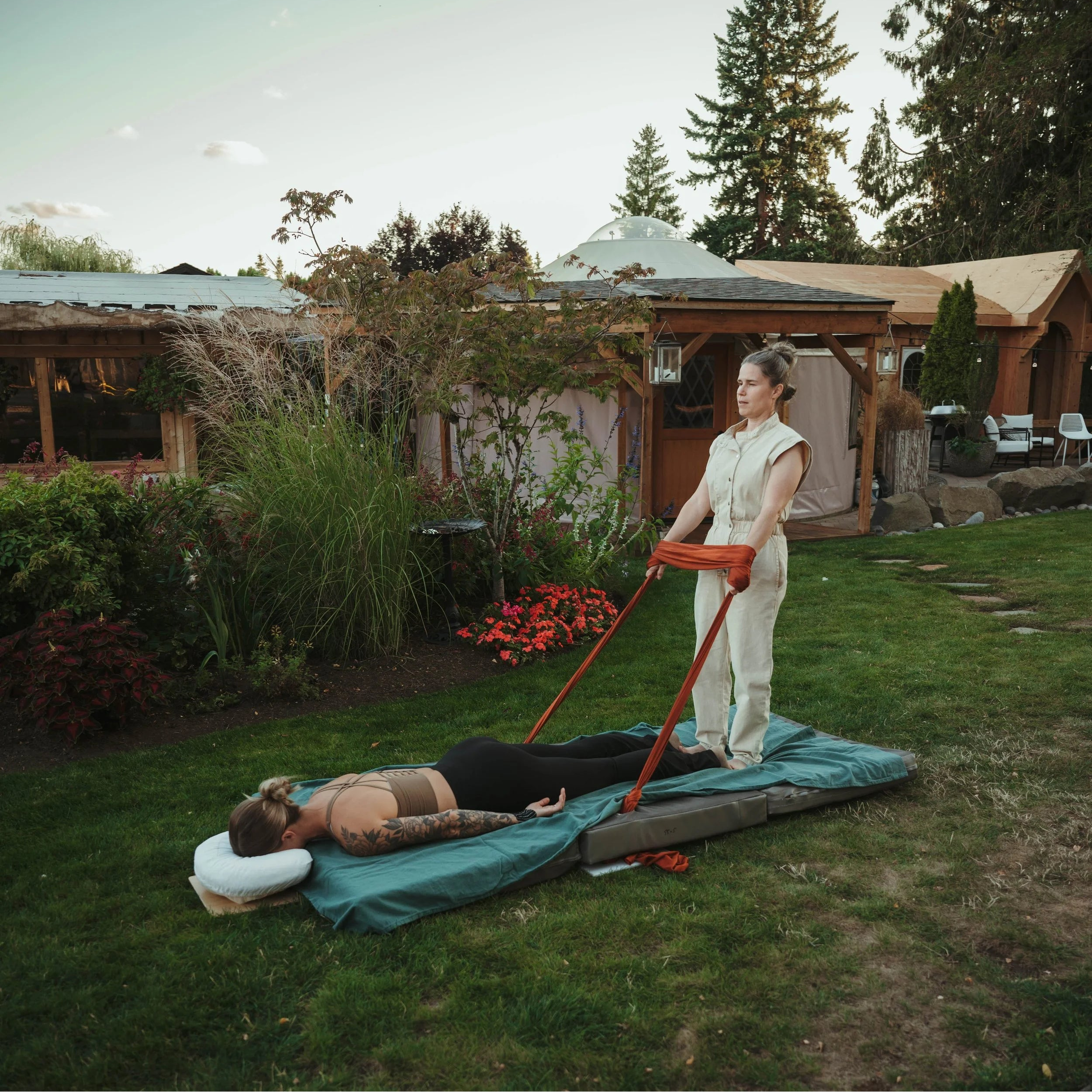 Person lying on a mat outdoors with another person using a tool on them, surrounded by greenery and a building.